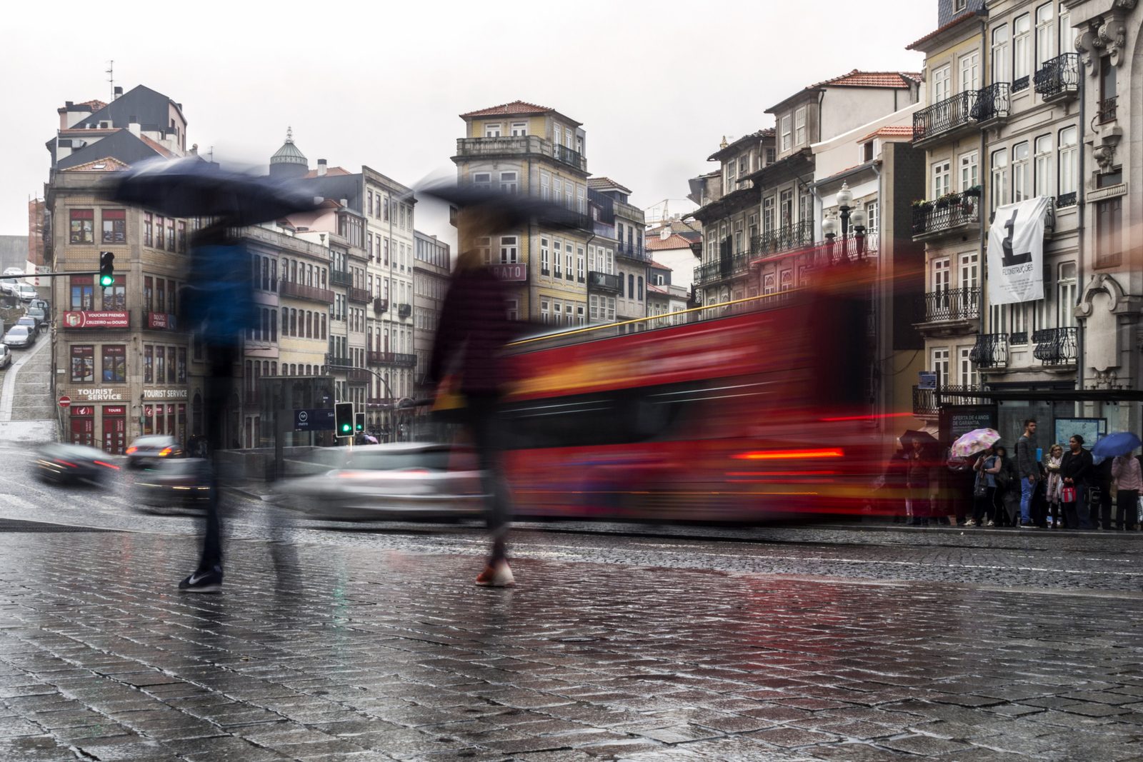 Deszczowa ulica w Porto: dwie rozmyte w ruchu postacie pod parasolami przechodzą po mokrym bruku, za nimi czerwony autobus i kolorowe kamienice