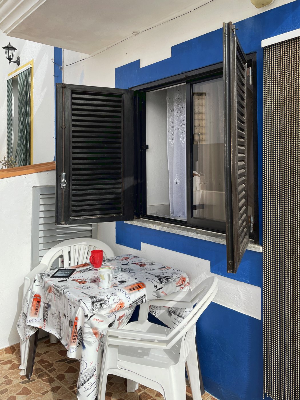 Patio domku w Algarve: plastikowy stolik z obrusem i krzesła pod oknem z otwartymi okiennicami, ściany w białe i niebieskie pasy