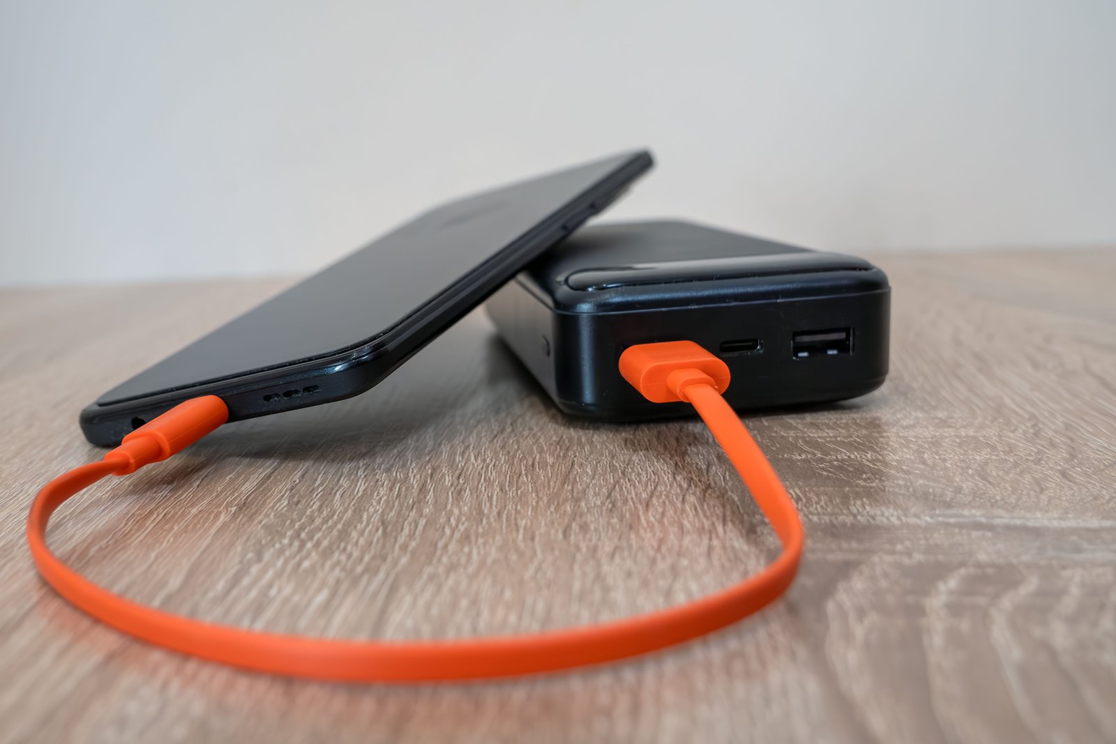 Telefon podłączony pomarańczowym kablem do powerbanku, oba leżą na drewnianym stole